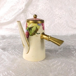 Op de afbeelding: Crèmekleurige keramische koffiepot met een gouden handvat en tuit. De pot heeft een deksel met een gouden knop en is versierd met een geschilderd ontwerp van druiven en bladeren in rood, groen en paars.