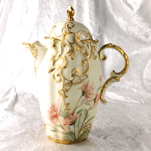 May include: An antique white porcelain coffee pot with a lid, adorned with gold filigree and a floral design. The pot features a curved handle and a spout. Pink and red flowers are painted on the front of the pot.