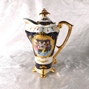 May include: A vintage porcelain coffee pot with a gold handle and accents. The pot features a navy blue and white design with a detailed scene of people dining. The lid is topped with a gold finial, and the base has three gold feet.