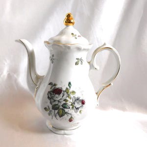 Puede incluir: Cafetera de porcelana blanca con tapa y remate dorado. La cafetera presenta un diseño floral con rosas rojas y blancas y hojas verdes. Los bordes dorados acentúan el asa, el pico y la tapa.