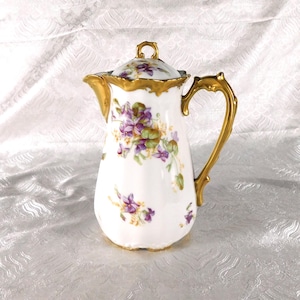 May include: White porcelain coffee pot with a lid, featuring a gold handle and trim. The pot is decorated with purple and green floral patterns. The handle and trim have a gold finish.