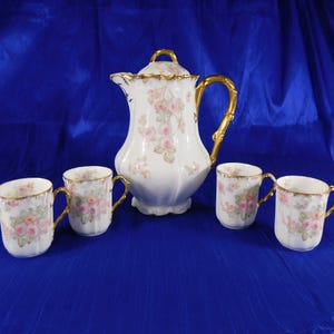 May include: A white porcelain coffee set featuring a tall, lidded pitcher and four matching mugs. Each piece is decorated with soft pink and orange floral patterns, scalloped edges, and ornate gold-painted handles and rim accents.