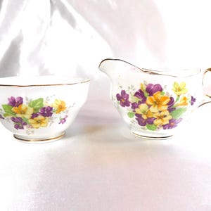 May include: A white porcelain creamer and sugar bowl set. Both pieces feature a floral design with purple, yellow, and green flowers. The rims and handles are trimmed with gold.