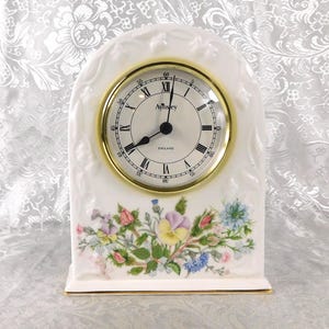 May include: A white porcelain mantel clock with a gold-tone bezel and Roman numeral clock face. The clock features a floral design at the base and the text "Aynsley England".