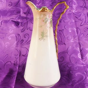 May include: A porcelain pitcher with a tapered body, featuring a soft cream and white gradient finish. The exterior is decorated with delicate hand-painted pink floral motifs and green leaves. It has a gold-trimmed rim and a wavy gold handle.