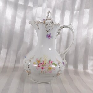 Puede incluir: Jarra de porcelana blanca con un diseño floral en rosa, amarillo y morado. La jarra tiene un asa curva y una tapa decorativa con un pequeño lazo. Los bordes y los detalles florales están adornados con ribetes dorados. Una pieza decorativa vintage.