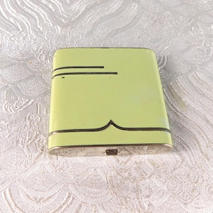 May include: A rectangular, pale lime green metal cigarette case featuring thin, dark horizontal lines and a central curved decorative accent. The case has a metallic edge and a small latch mechanism visible on the bottom side of the object.