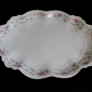 May include: Oval white porcelain serving platter with a scalloped edge. The rim is decorated with a delicate floral pattern of pink roses and green leaves. The platter has a smooth, glossy surface.