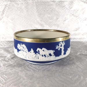 May include: A deep blue ceramic bowl featuring white relief carvings of horses, figures, and trees around the exterior. The bowl has a smooth, metallic gold-toned rim and a white interior, with a decorative white pattern along the base edge.