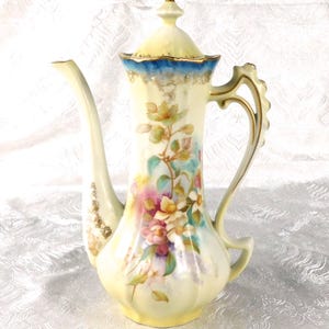 Könnte beinhalten: Eine hohe Kaffeekanne aus Porzellan mit cremefarbenem Körper und handgemaltem Blumenmotiv in Rosa-, Gelb- und Grüntönen. Der Rand ist mit einem leuchtend blauen Band und Goldverzierungen versehen, ergänzt durch einen passenden Deckel mit Goldknauf.