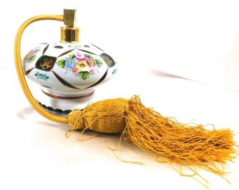 Könnte beinhalten: Eine Vintage-Parfümflasche mit weißer Keramikbasis, verziert mit bunten Blumenmustern und goldenen Akzenten. Sie verfügt über einen goldfarbenen Zerstäuber und eine lange, dekorative goldene Quaste. Die Flasche ist ein klassischer Dekorationsartikel.