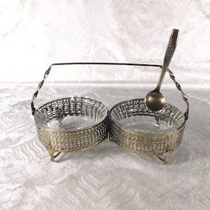 May include: A double serving dish set featuring two clear glass bowls encased in ornate silver-toned metal holders with a woven pattern. The set includes a matching silver-toned metal handle and a single silver-toned metal spoon resting inside.