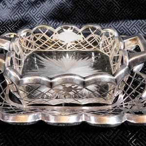 May include: A silver serving dish with a decorative, geometric pattern. The dish has a square, clear glass insert with a starburst design.