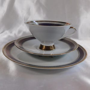 May include: A white teacup and saucer set with gold trim and a dark blue and gold patterned border. The teacup sits on a matching saucer and a larger plate, all on a white background.