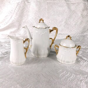Puede incluir: Un juego de té de porcelana blanca que consta de una tetera, una lechera y un azucarero con tapa. Cada pieza presenta un diseño de borde festoneado y está decorada con asas, remates y bordes de color dorado en los bordes y picos.