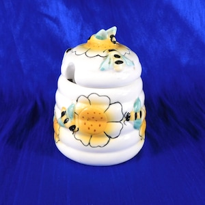 Pode incluir: Um pote de mel de cerâmica branca com um formato canelado inspirado numa colmeia. O exterior apresenta flores pintadas à mão em amarelo com centros cor de laranja e pequenas abelhas pretas e amarelas com asas azul-claras.