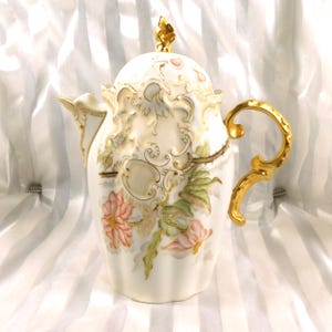 May include: An ornate, cream-colored porcelain teapot with a gold handle and accents. The teapot features a floral design with pink and green flowers, and a decorative lid with a gold finial. The teapot is a classic piece of tableware.