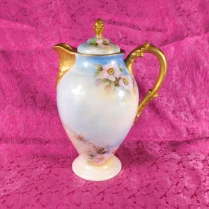 Op de afbeelding: Een porseleinen koffiepot met een handgeschilderd bloemmotief met roze bloemen en groene bladeren tegen een zachte blauwe en witte achtergrond. De pot heeft een goudkleurig handvat, een tuit en een knop op het deksel.