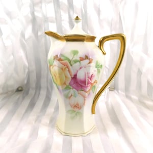 Può includere: Una caffettiera vintage in porcellana con coperchio, con un motivo floreale di rose rosa, gialle e pesca. La caffettiera ha un manico e finiture dorate e uno sfondo bianco.