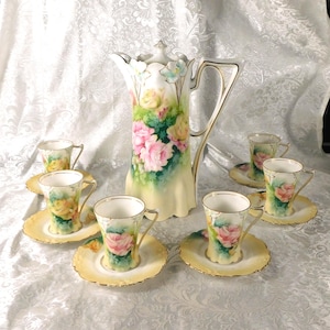May include: A vintage porcelain tea set featuring a tall pitcher and six teacups with saucers. The set is decorated with hand-painted pink and yellow roses on a cream-colored background. The pitcher has a gold handle, and the cups have gold trim.