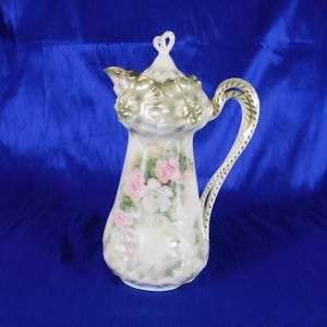 May include: A porcelain pitcher featuring a white base with a hand-painted floral motif of pink and white roses. The vessel includes a removable lid with a decorative finial, a ruffled rim, and a curved handle with gold-toned trim accents.