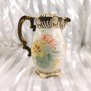May include: Antique porcelain pitcher with a floral design featuring yellow and peach flowers, green leaves, and gold accents. The pitcher has a decorative handle and base, and a matching lid. The overall design is elegant and vintage.