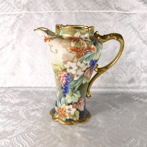 May include: A porcelain jug featuring a scalloped rim and a gold-painted handle. The body is decorated with hand-painted floral motifs, including white blossoms, blue grapes, and green leaves, set against a cream and blue gradient background.