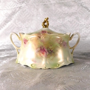 May include: A vintage porcelain serving dish with a lid, featuring a hand-painted floral design of pink roses and green leaves. The dish has a pearlescent finish, gold trim, and a gold-coloured handle on the lid.
