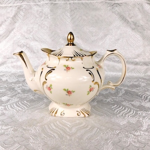 Puede incluir: Una tetera de porcelana color crema con un delicado patrón de pequeñas rosas rosadas con hojas verdes. La tetera presenta detalles en oro a lo largo del pico, el asa, la base y la tapa, la cual está rematada con un pequeño remate dorado.