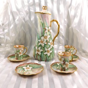 May include: A hand-painted porcelain coffee set featuring a tall, slender coffee pot with a gold handle and lid, and four cups and saucers. The set is decorated with white daffodils and green foliage against a soft pink and green background.