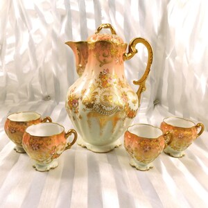 May include: A vintage porcelain tea set featuring a large peach-colored coffee pot with a lid and four matching teacups. The set is decorated with gold accents and floral patterns, creating an elegant and antique aesthetic.