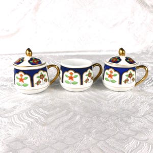 May include: Three porcelain cups, two with matching lids, featuring a white base with a deep blue scalloped border. Each cup is decorated with small orange and red floral motifs and green leaves. The handles and lid finials are gold-toned.