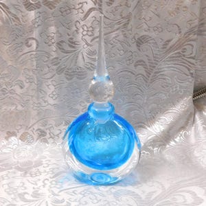 Può includere: Un flacone di profumo in vetro trasparente con un interno blu vibrante e un tappo trasparente a punta. Il flacone ha una forma arrotondata con una base bulbosa e un lungo tappo sottile. Il colore blu è concentrato al centro.