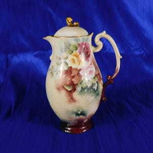Può includere: Una brocca in porcellana con coperchio caratterizzata da un motivo floreale dipinto a mano con rose rosse, gialle e rosa su sfondo color crema. La brocca presenta un pomello dorato sul coperchio, un manico curvo e una base bordeaux.
