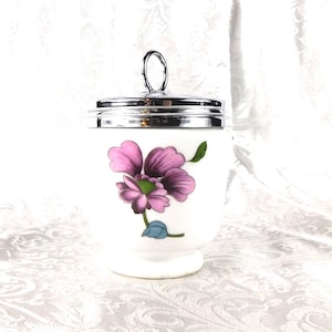 May include: White ceramic egg cup with a silver-tone lid and handle. The egg cup features a floral design with pink and purple petals, green leaves, and a blue accent. A decorative kitchenware item.