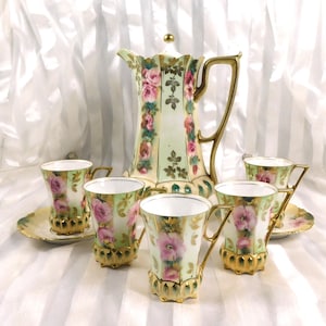 May include: A vintage porcelain tea set with a tall coffee pot and five cups. The set is decorated with pink roses, gold trim, and a light green background. The cups have a unique, ornate base. The set is a classic design.