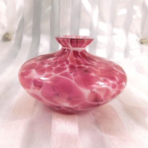 May include: A round, pink glass vase with a speckled pattern. The vase has a wide, bulbous body and a narrow neck. The pink color is interspersed with white, creating a unique, artistic design. The vase is likely used for decorative purposes.