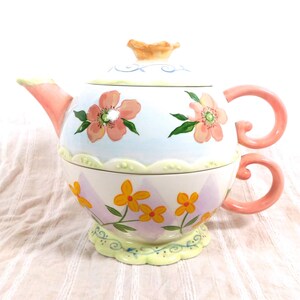 May include: A decorative ceramic teapot and teacup set. The teapot is white with pink and yellow floral designs, a peach-colored spout, and a matching handle. The teacup is stacked below the teapot, with a matching handle.