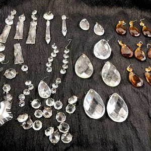 May include: Assortment of clear and amber crystal chandelier prisms and beads on a black fabric background. Various shapes include teardrops, faceted pendants, and round beads. Some have gold-colored metal accents.