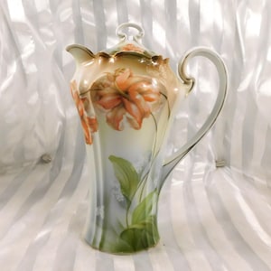 May include: A tall, antique porcelain coffee pot with a lid, featuring a hand-painted floral design. The pot has a cream-colored base with orange and peach-colored flowers and green leaves. The handle and rim are accented with gold trim.
