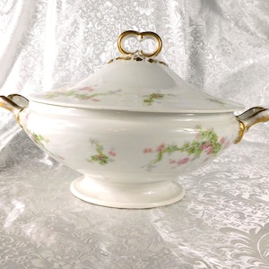 Op de afbeelding: Witte porseleinen serveerschaal met deksel, versierd met roze bloemmotieven en gouden details. De schaal heeft een voet en gouden handvatten. Een klassiek stuk voor het serveren van soepen of stoofschotels. Ongeveer 25 cm in diameter.