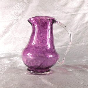 May include: A handcrafted, purple glass pitcher with a clear, curved handle. The pitcher has a bulbous body and a flared rim. The glass has a speckled pattern, and the base has a silver rim. This decorative pitcher is suitable for serving beverages.