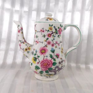 May include: White ceramic coffee pot with a lid, featuring a floral design with pink, yellow, and blue flowers. The handle and rim have green accents. A decorative piece for serving coffee or tea.