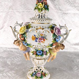 May include: A white porcelain urn with a lid, featuring hand-painted floral bouquets, gold trim, and two cherub figures attached to the sides. The piece is decorated with three-dimensional ceramic flowers in yellow, purple, blue, and red.
