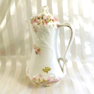 Może przedstawiać: Biały porcelanowy dzbanek do kawy z pokrywką, ozdobiony delikatnymi różowymi i żółtymi wzorami kwiatowymi oraz złotymi zdobieniami. Uchwyt jest wdzięcznie zakrzywiony, a dzbanek ma smukły, elegancki kształt. Klasyczny element do serwowania napojów.