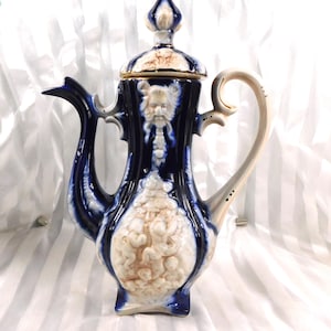 Puede incluir: Una cafetera de cerámica vintage con un esmalte azul oscuro y detalles en relieve ornamentados. La cafetera presenta un asa y detalles en color crema, una tapa decorativa y un pico. El diseño incluye una cara y otros elementos clásicos.