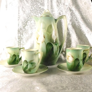 Op de afbeelding: Een vintage theeservies met een hoge, sierlijke theepot en vier kopjes met schotels. De set is geschilderd in groen en wit, met een bloemmotief. De theepot heeft een gouden handvat en tuit. De kopjes hebben een gouden rand.