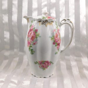 Puede incluir: Cafetera de porcelana blanca con tapa, adornada con rosas rosas y hojas verdes. La jarra tiene un borde festoneado y un asa dorada. Una pieza decorativa para servir bebidas.