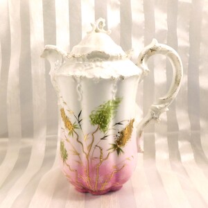 May include: A white porcelain coffee pot with a lid, handle, and spout. The pot features a pink ombre base, white body, and gold and green floral design. The handle and rim have gold accents.