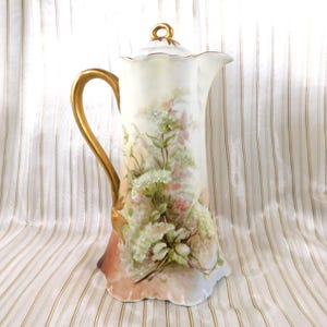 Puede incluir: Una jarra alta de porcelana antigua con asa y detalles dorados. La jarra presenta diseños florales pintados a mano en blanco, rosa y verde. El asa y la parte superior son doradas. Un objeto decorativo para el hogar, de unos 25 cm de altura.